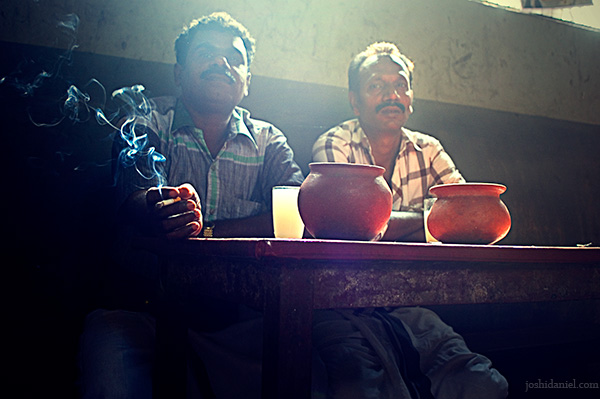 Two men with glasses of toddy behind a swirl of cigarette smoke in a kallu shap in Kozhikode, Kerala