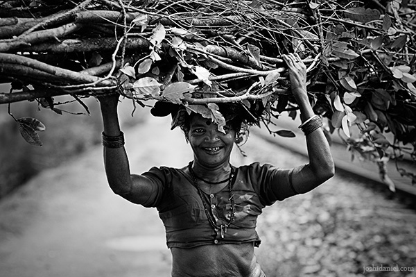 Black and white photograph of a smiling woman carrying fire wood on her head in Kumta, Karnataka