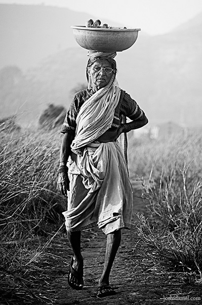 Black and white photograph of a an old woman carrying a load on her head in Pune