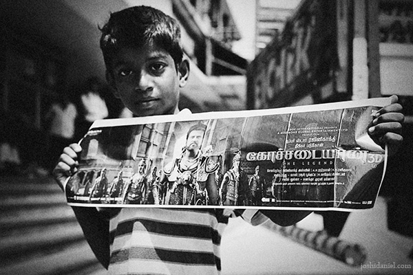 Black and white portrait of a boy holding a Kochadaiiyaan movie poster starring Rajinikanth at Udhayam theatre in Chennai