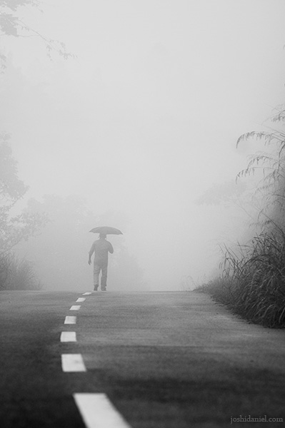 Black and white conceptual photograph of a man holding an umbrella walking on a foggy road in Ponmudi, Kerala